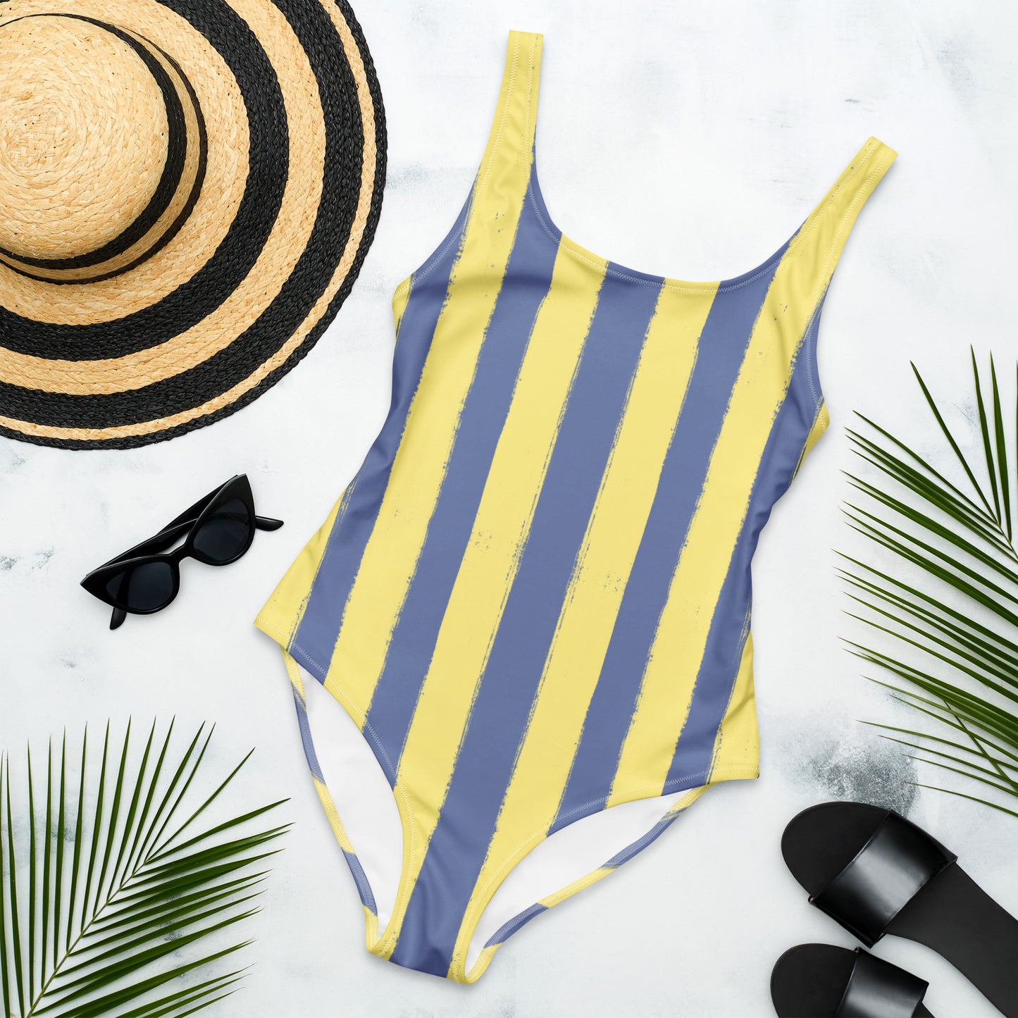 Flat lay of Panther & Palm butter yellow and blue one-piece swimsuit with Riviera-style stripes, styled with beach essentials for effortless vacation dressing.