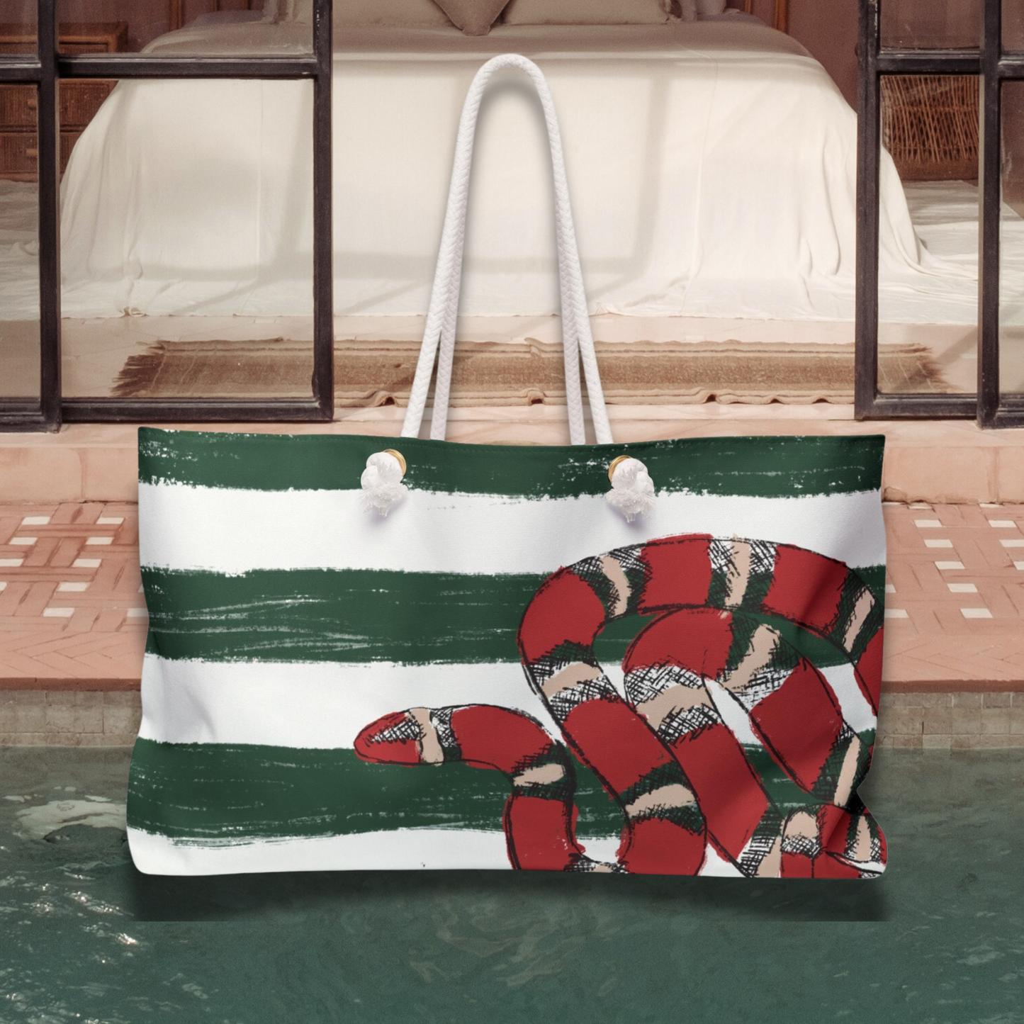 Snake Stripe Weekender Bag staged poolside with its reflection on the water, emphasizing the vibrant green and cream stripes with red snake detail.