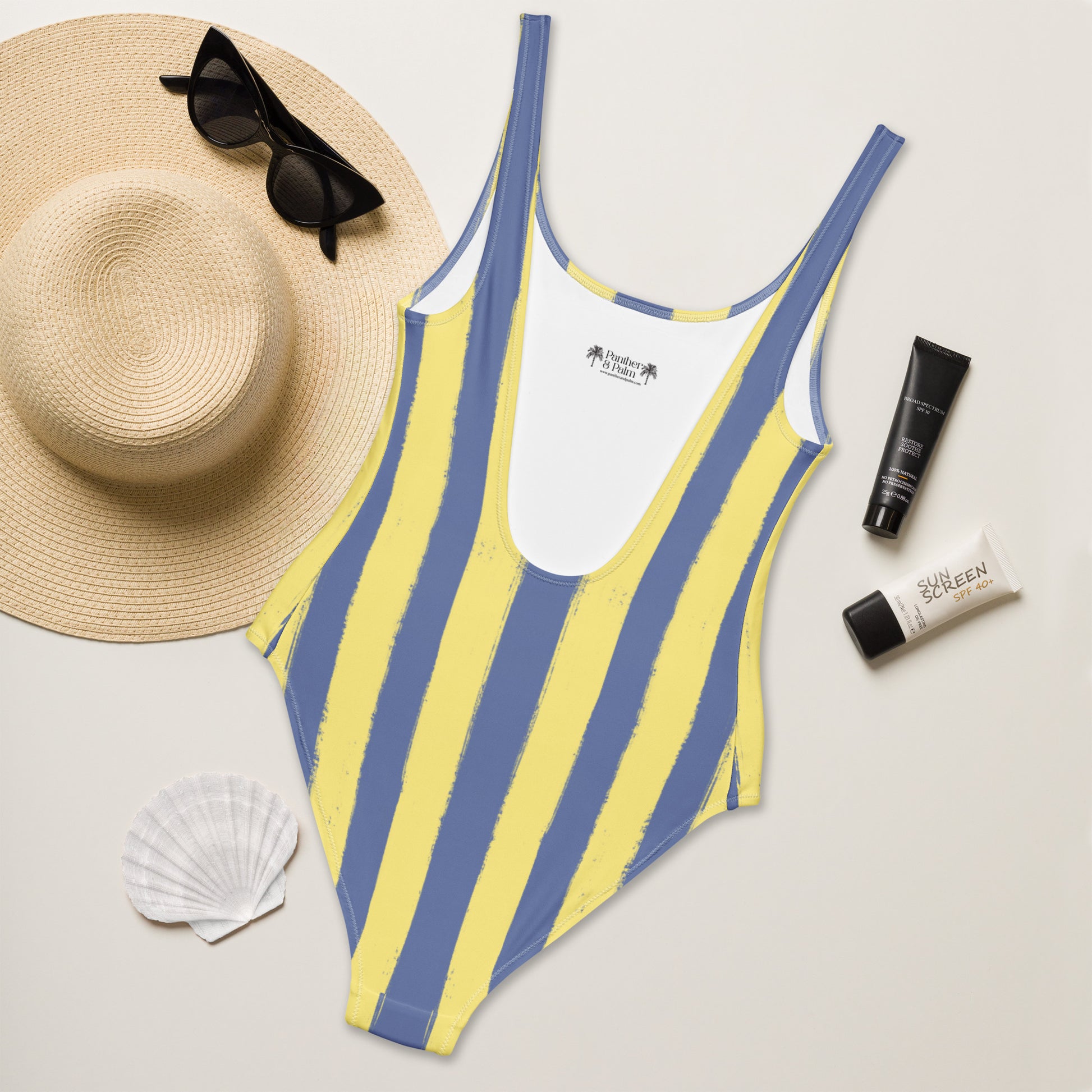 Flat lay of Panther & Palm butter yellow and blue one-piece swimsuit with Riviera-style stripes, styled with beach essentials for effortless vacation dressing.